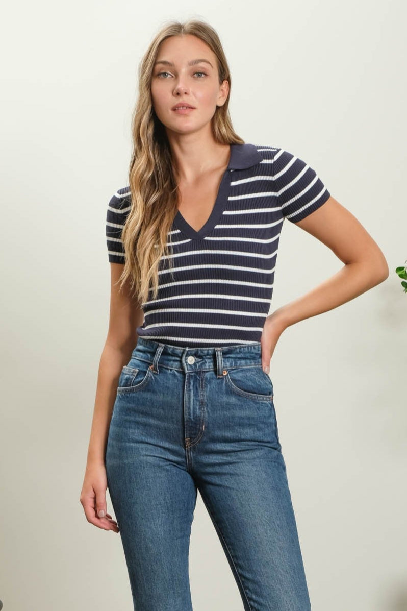 Woman wearing a striped shirt and blue jeans standing in a room with a plant and chair.