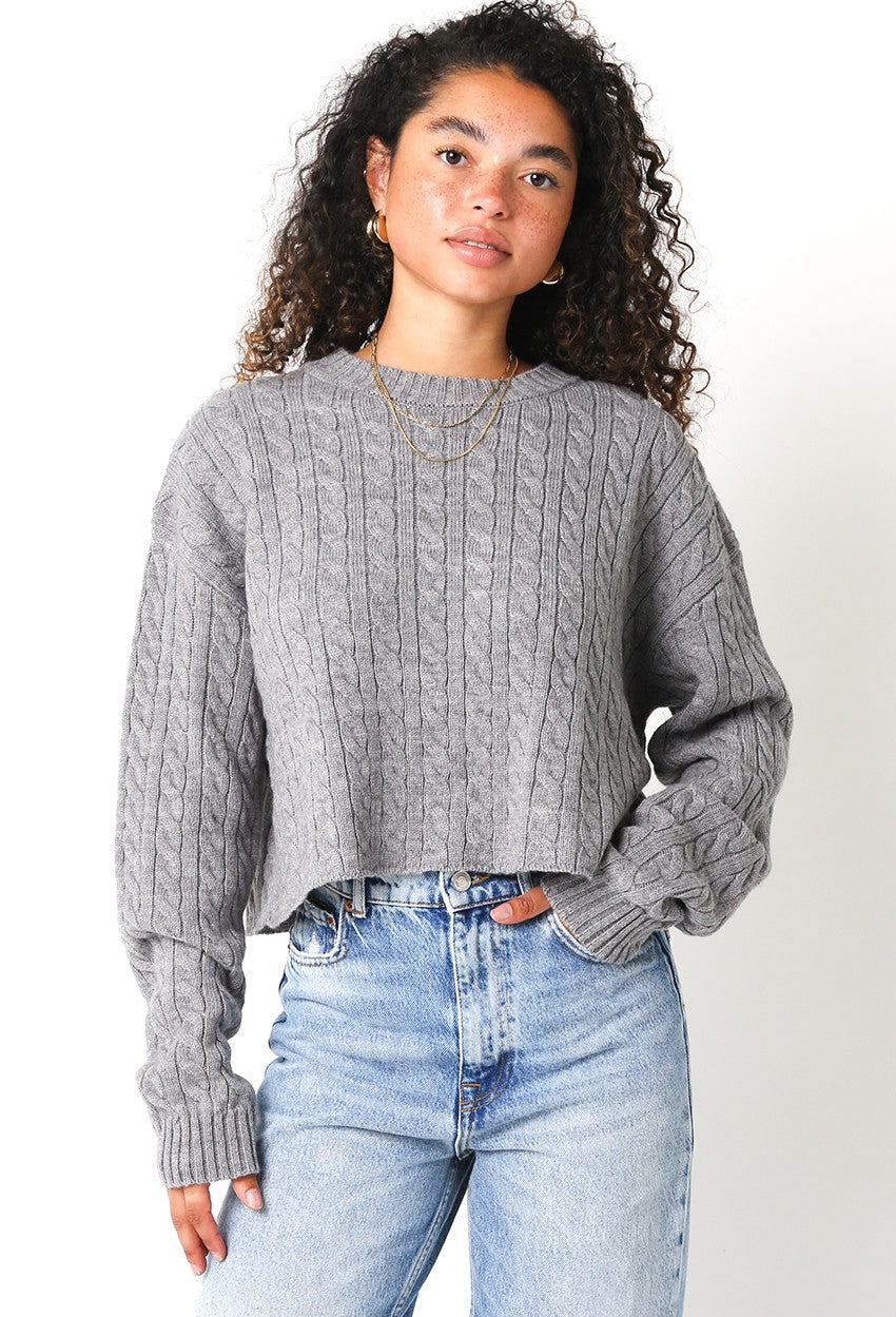 Woman wearing a gray cable knit sweater and light blue jeans on a white background