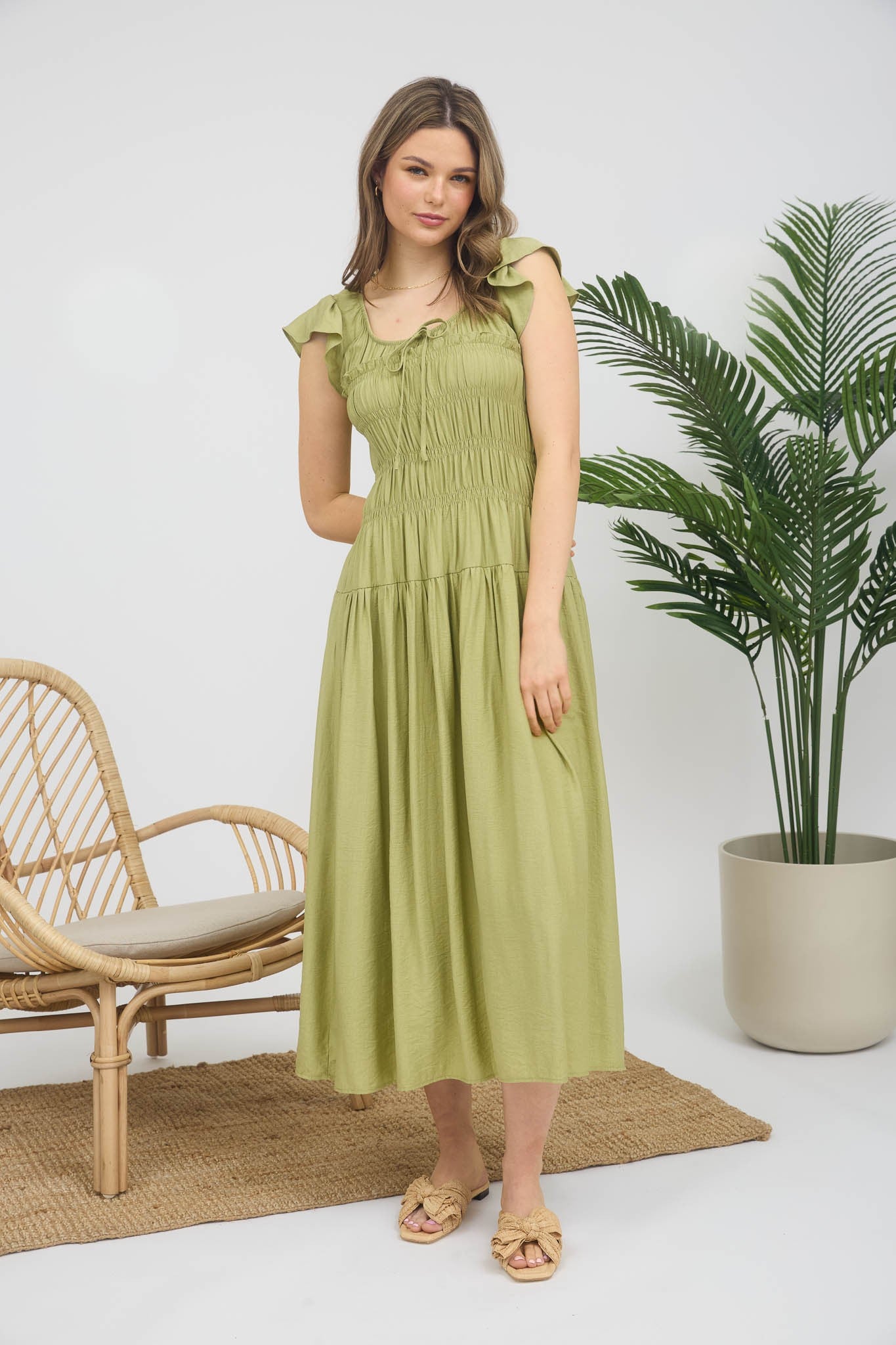 Woman in a green dress standing indoors with a plant and chair in the background.