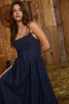 Woman in a dark blue dress standing in front of a wooden wall with a rustic feel.