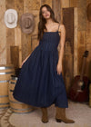 Woman in a blue dress standing in front of a wooden wall with hats and a guitar.