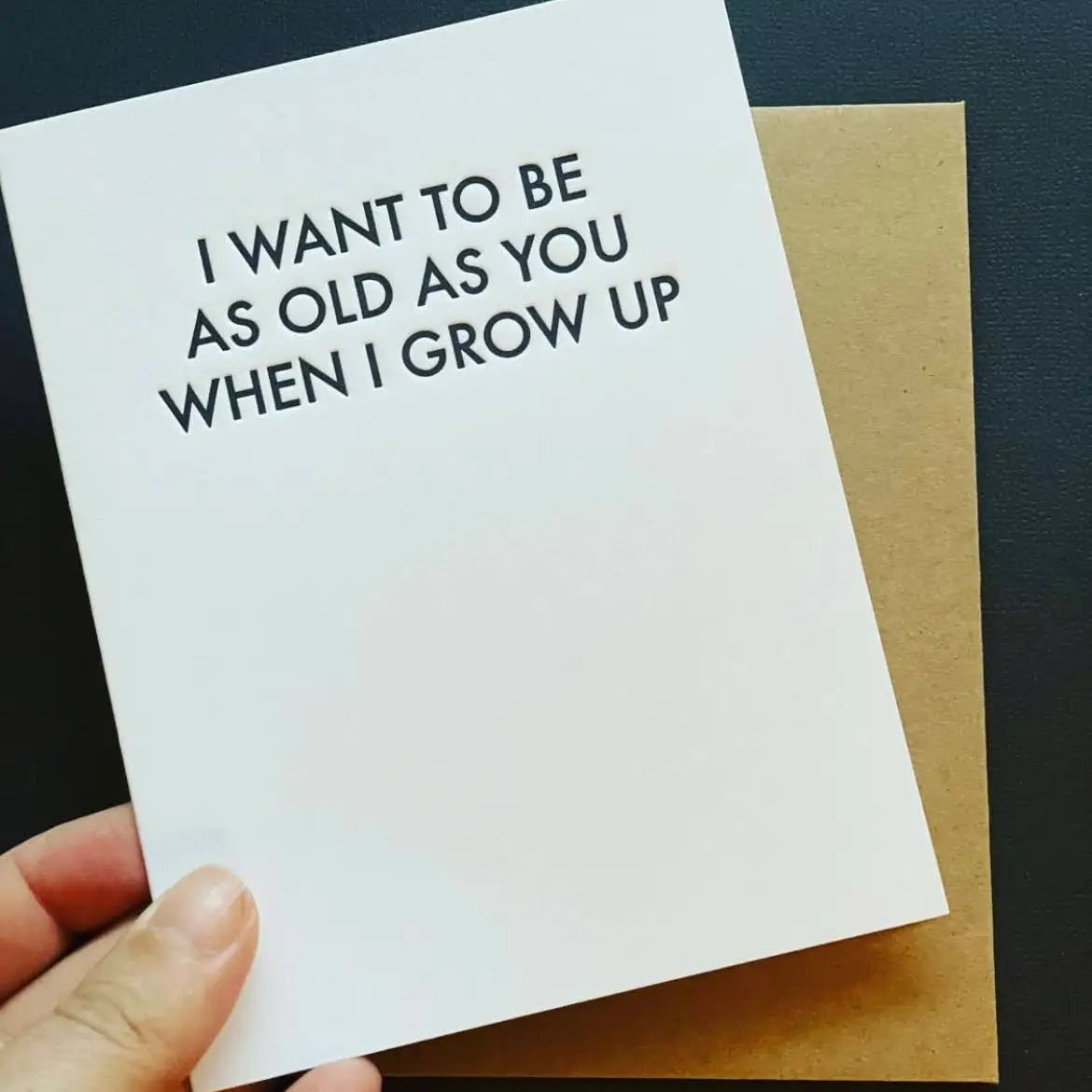 A hand holding a white A2 size birthday card with the text 'I WANT TO BE AS OLD AS YOU WHEN I GROW UP' printed on it. There is a brown envelope and a white sleeve in the background.