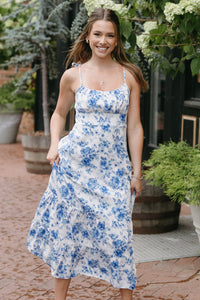 Woman in a floral dress standing outdoors with potted plants and a brick path.