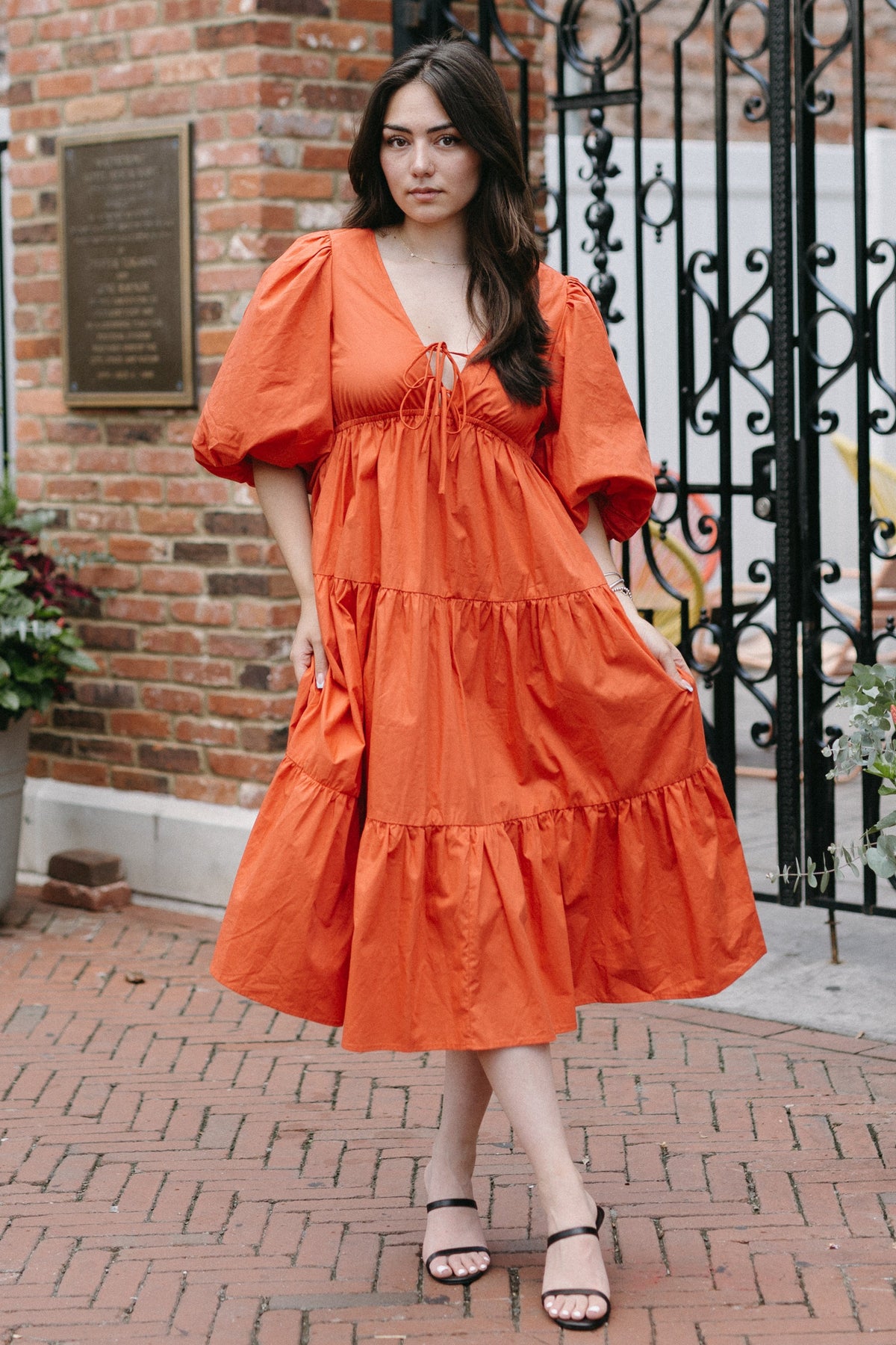 Woman in an orange dress standing in front of a brick building with decorative elements.
