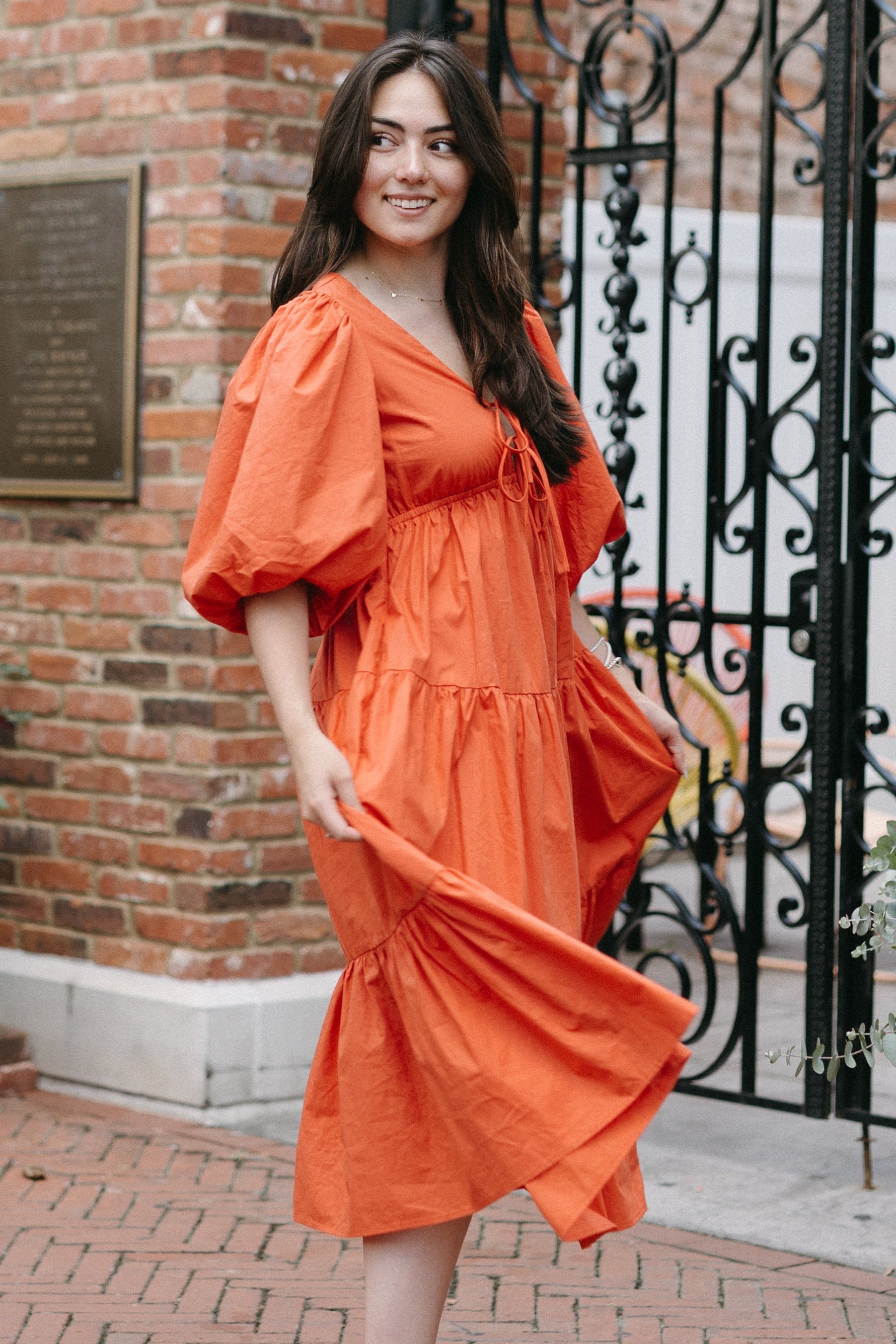 Woman in an orange dress standing in front of a brick wall with decorative elements.