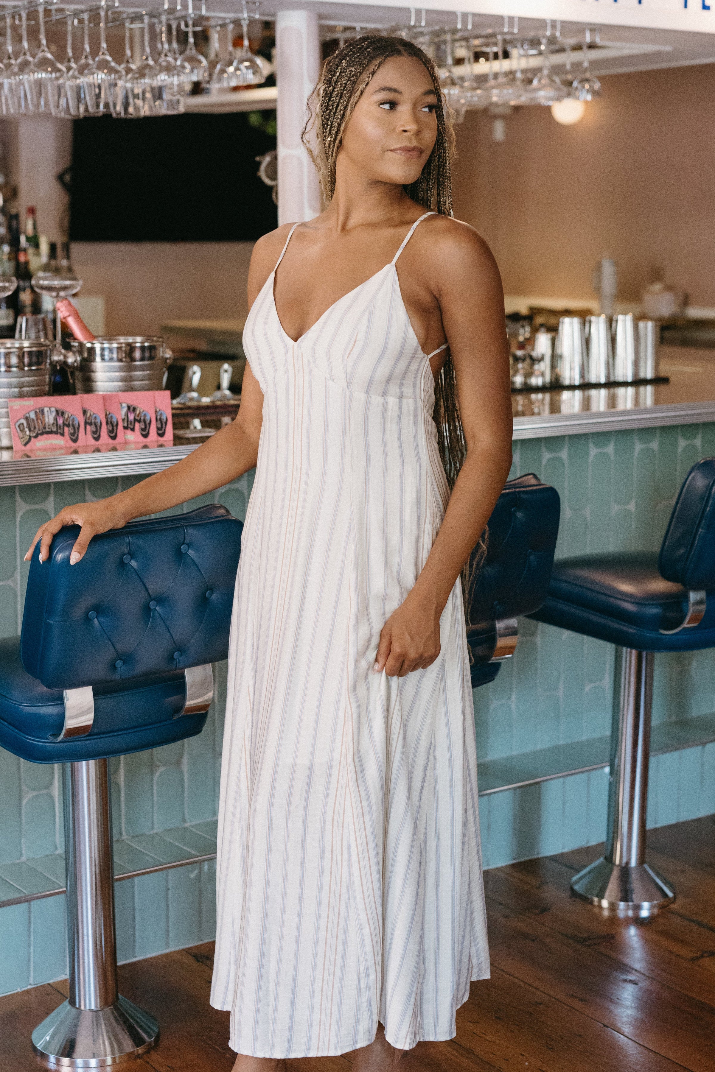 Woman in a white dress standing in a bar with blue stools and a counter.