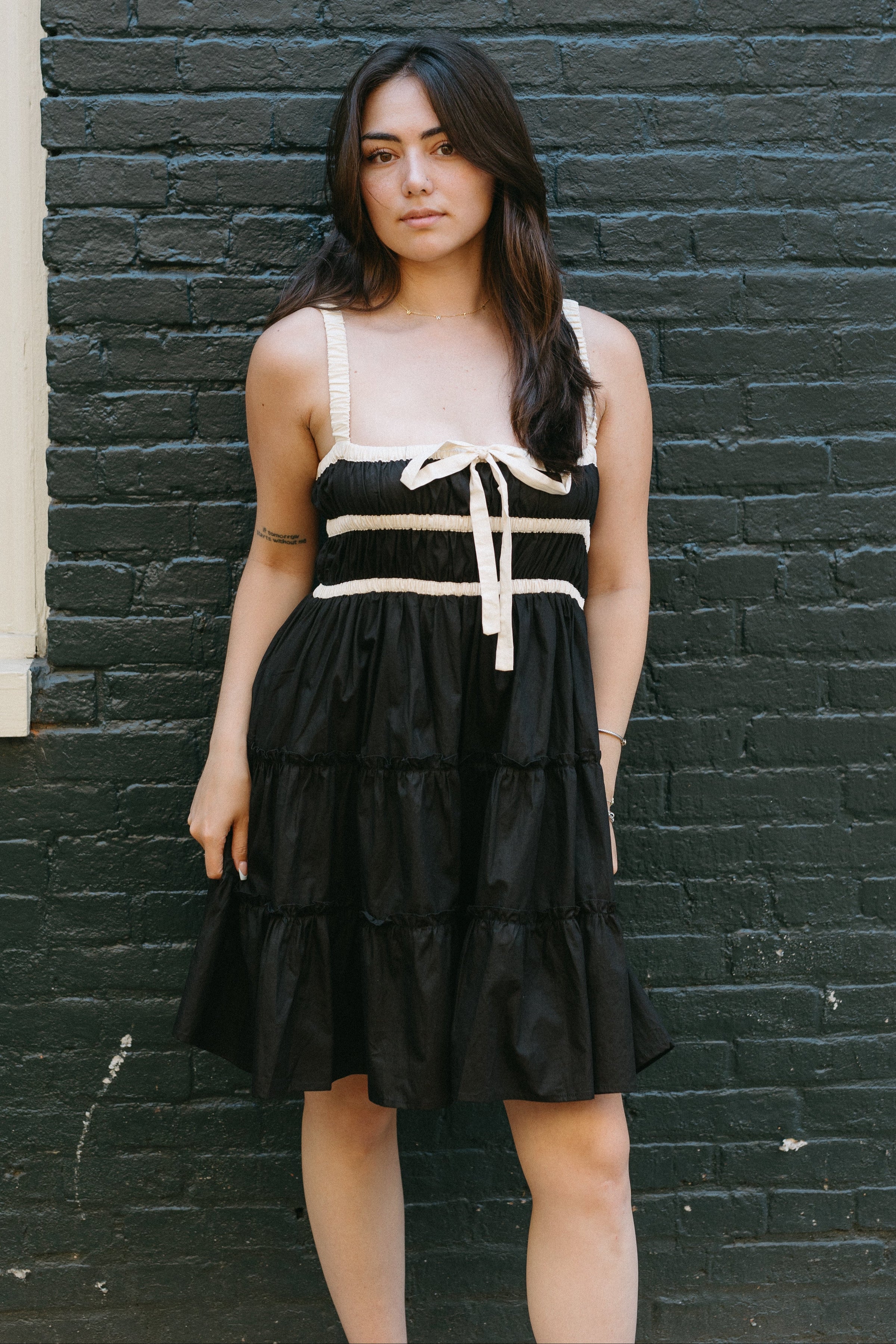 Woman wearing a black dress with white accents against a dark brick wall.