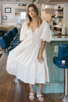 Woman in a white dress standing in a modern cafe with blue chairs and wooden floors.
