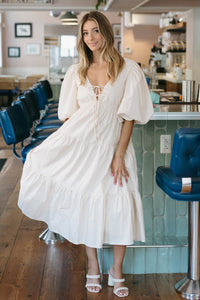 Woman in a white dress standing in a modern cafe with blue chairs and wooden floors.
