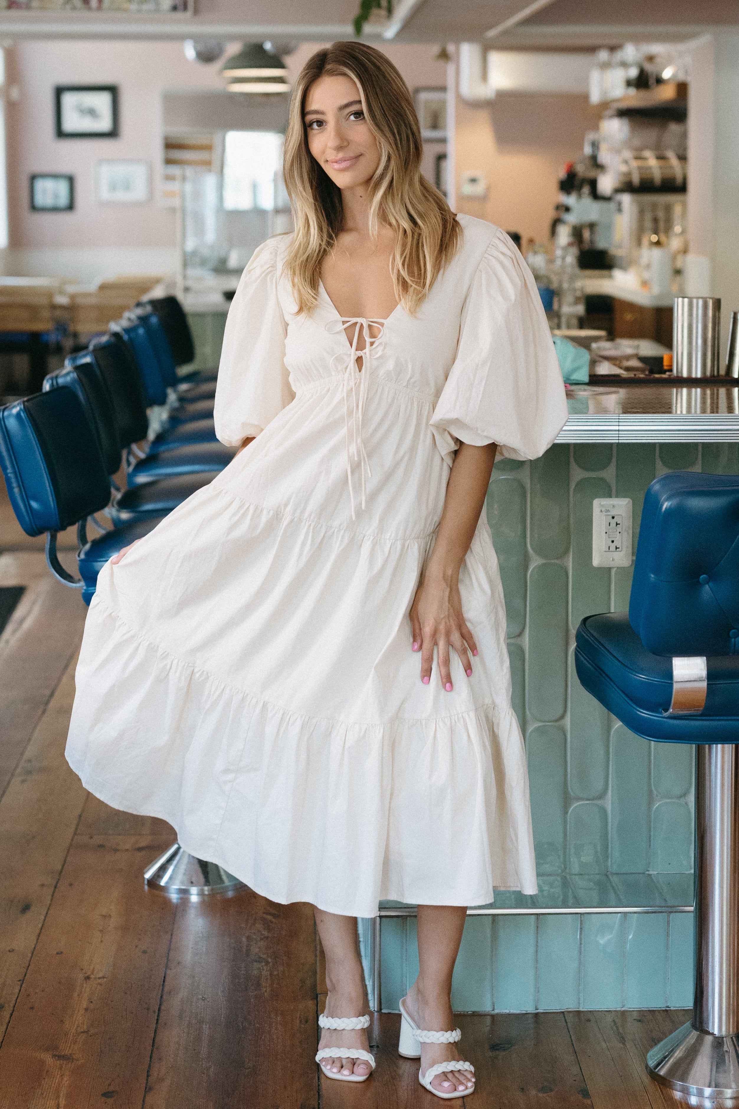 Woman in a white dress standing in a modern cafe with blue chairs and wooden floors.
