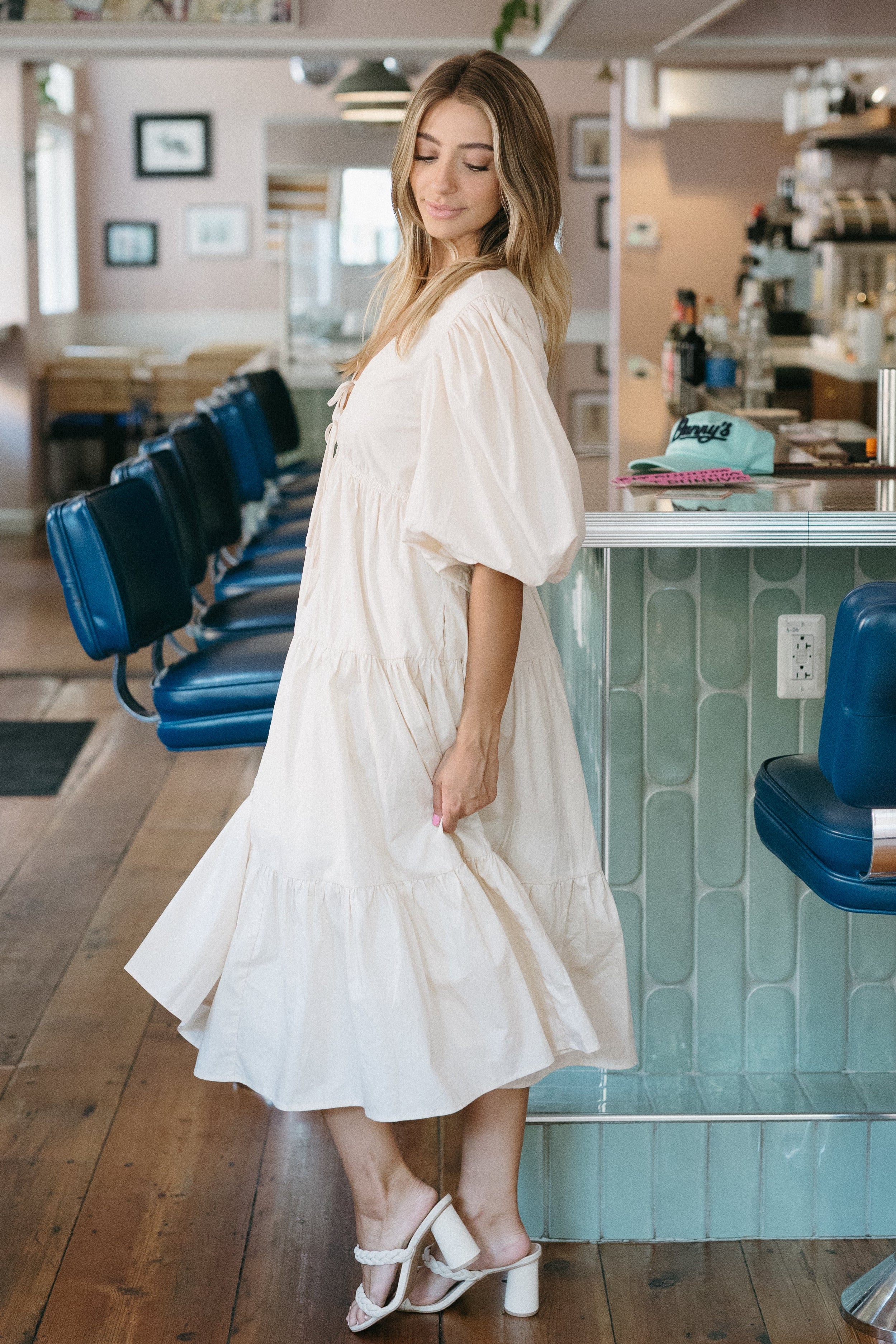 Woman in a white dress standing in a casual dining setting with blue chairs and a counter.