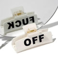 A black rectangular hair clip with the words 'FUCK OFF' written in white letters on the surface, placed against a white background with a mirror reflecting the clip.