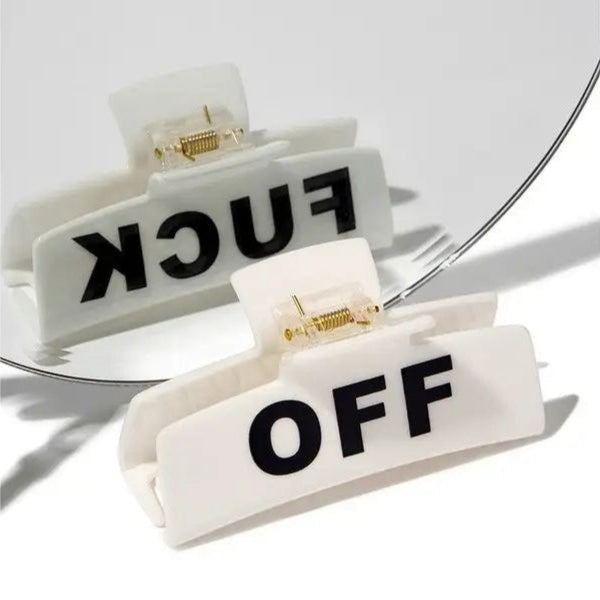 A black rectangular hair clip with the words 'FUCK OFF' written in white letters on the surface, placed against a white background with a mirror reflecting the clip.