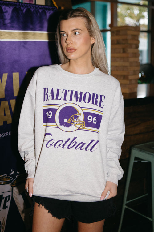 A woman wearing a white crewneck sweatshirt with 'Baltimore Football' text and the year '1996' on it, standing in front of a purple background with stadium seating visible in the distance.