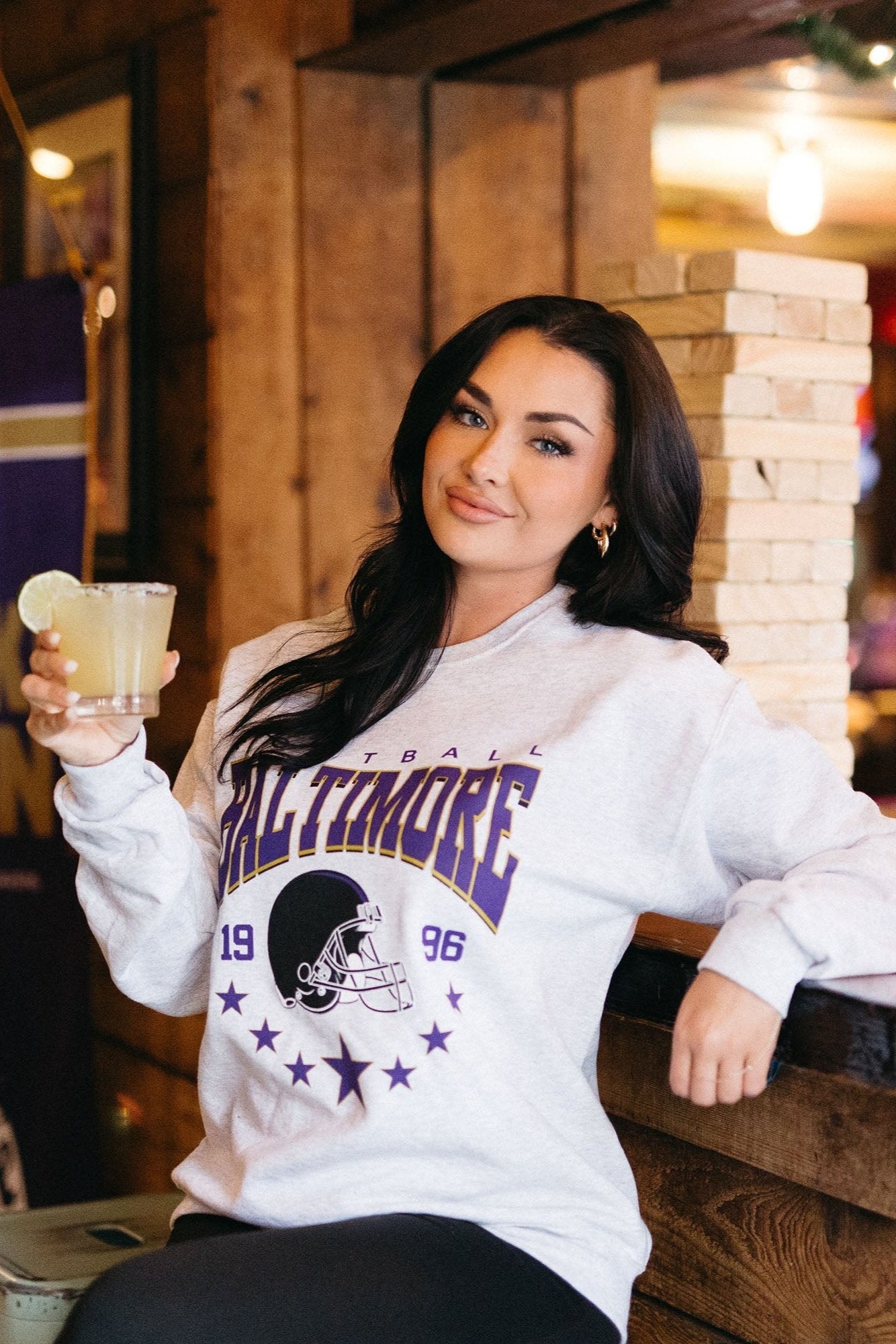 A woman sitting in a casual setting, wearing a white crewneck sweatshirt with the text 'Baltimore' and a football graphic on it.