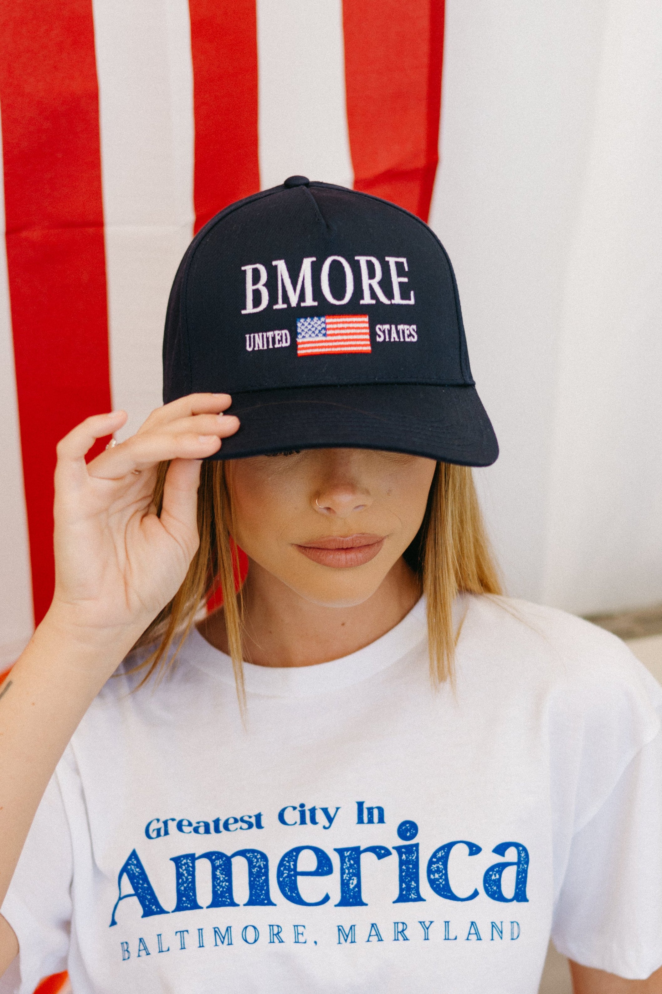 A person wearing a black snapback hat with 'BMORE' and an American flag design on it, sitting in front of a red and white striped background.