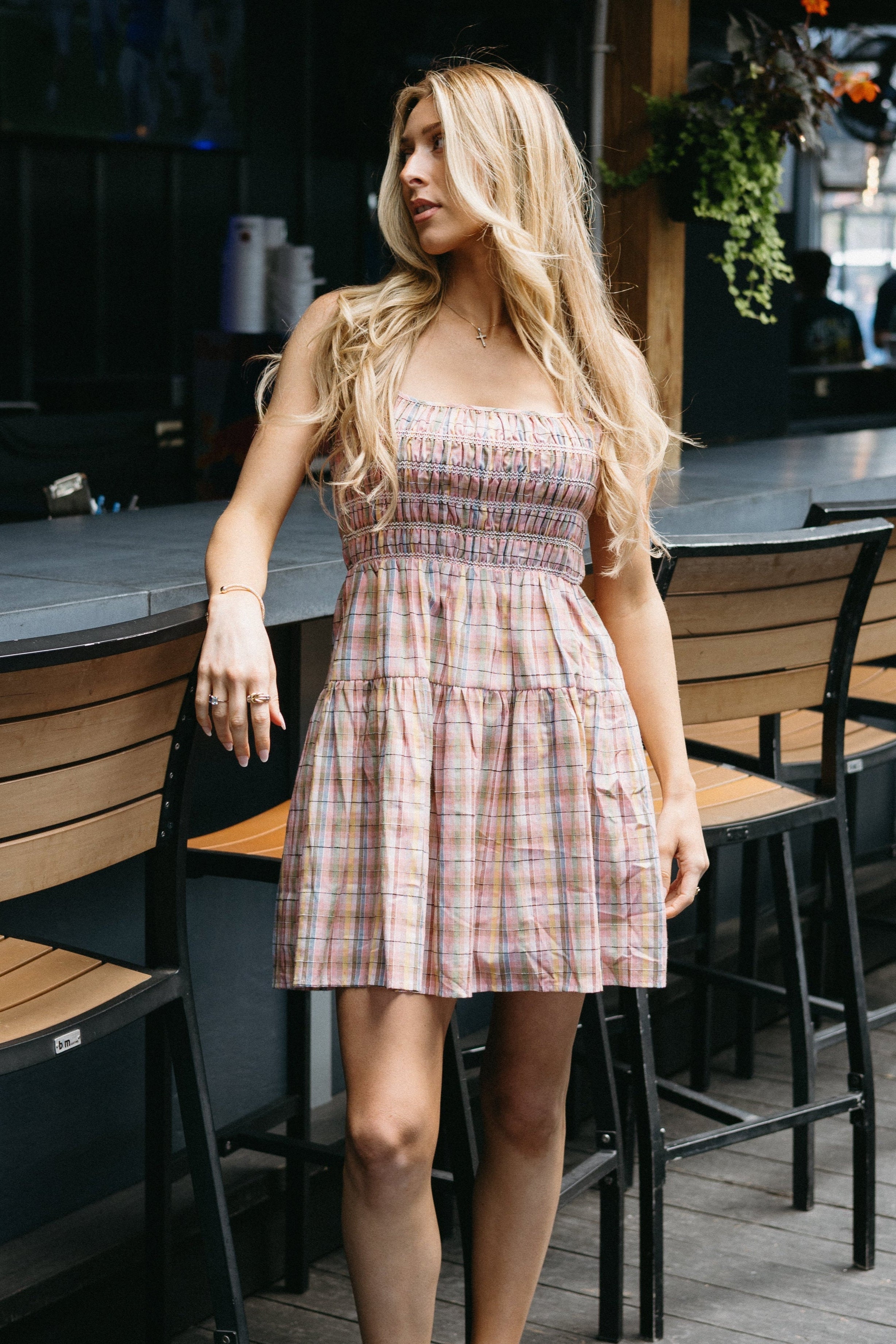 Woman in a plaid dress standing in an outdoor setting with tables and chairs.