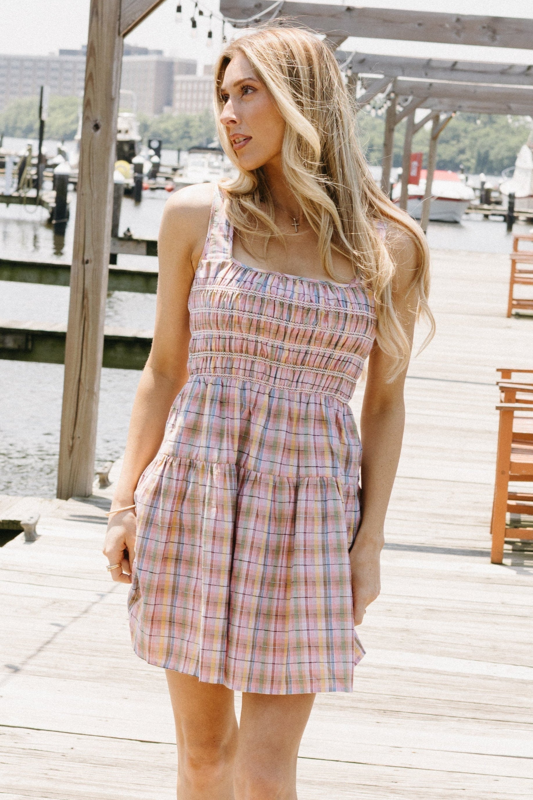 Woman wearing a plaid dress standing on a wooden dock by a waterfront.