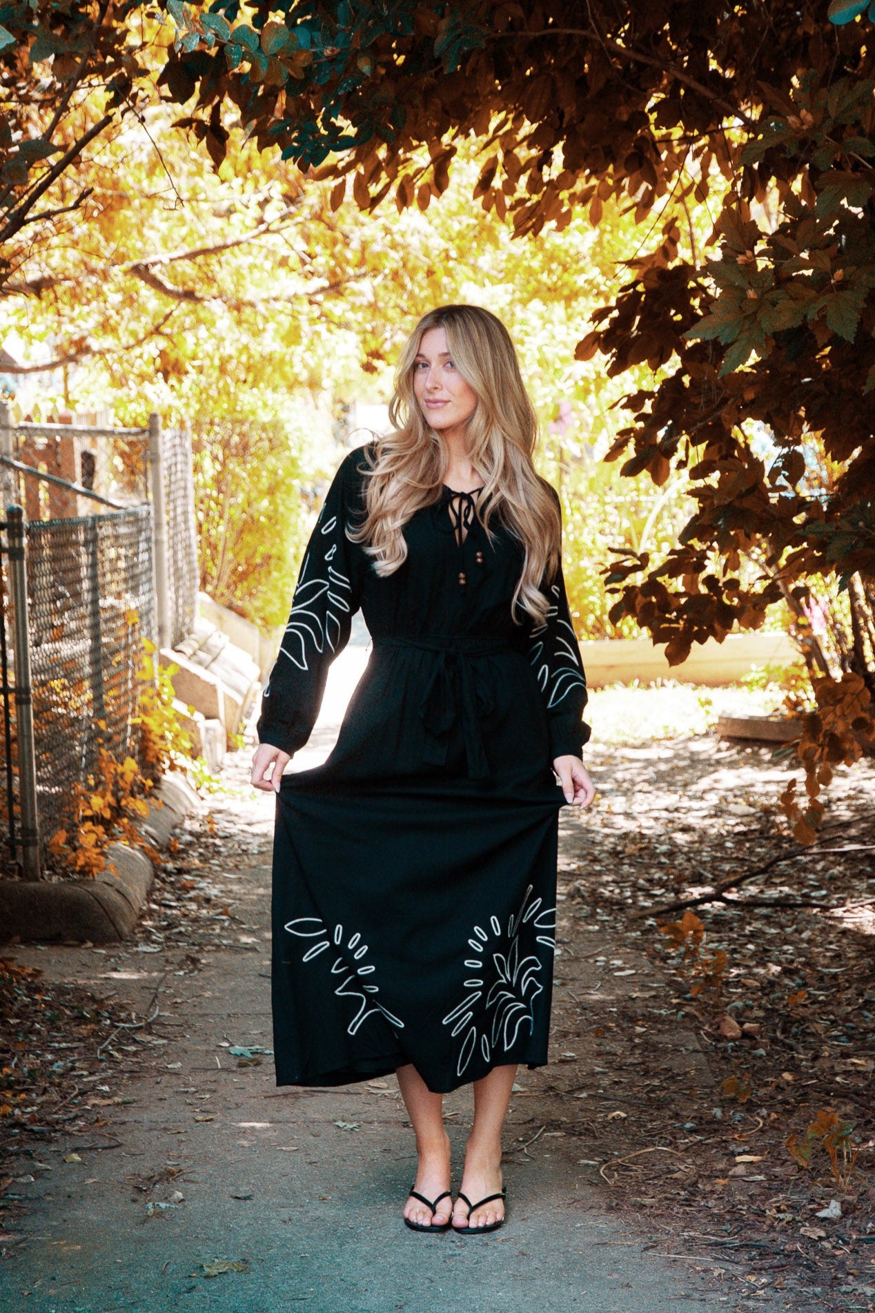Woman in a black dress with floral patterns standing in a sunlit forest path.