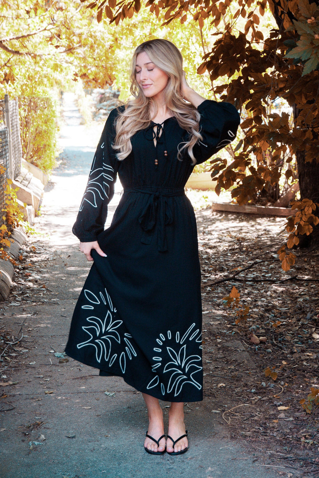 Woman in a black dress with white floral patterns standing under a tree with autumn leaves.