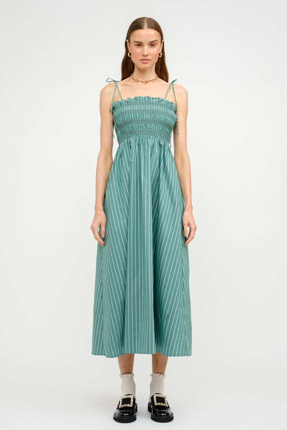 A model wearing a midi length green striped dress with shoulder ties and a smocked bodice.