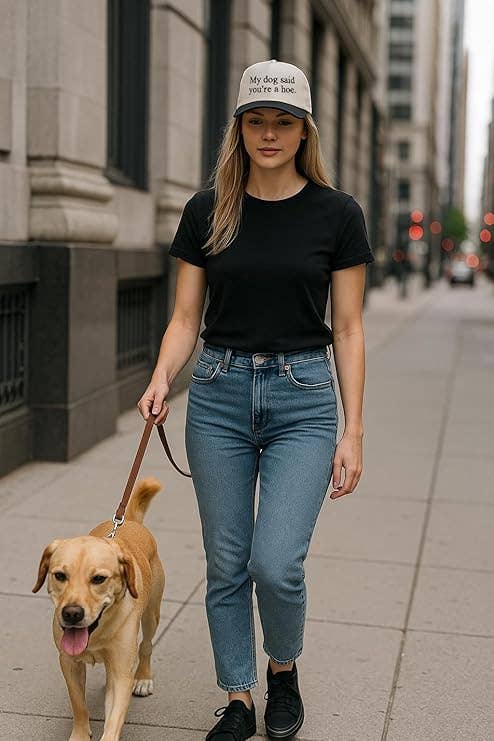Woman walking a dog on a city street wearing a black t-shirt, blue jeans, and a white cap.