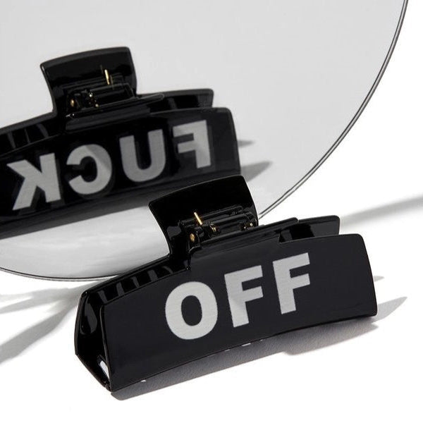 A black rectangular hair clip with the words 'FUCK OFF' written in white letters on the surface, placed against a white background with a mirror reflecting the clip.